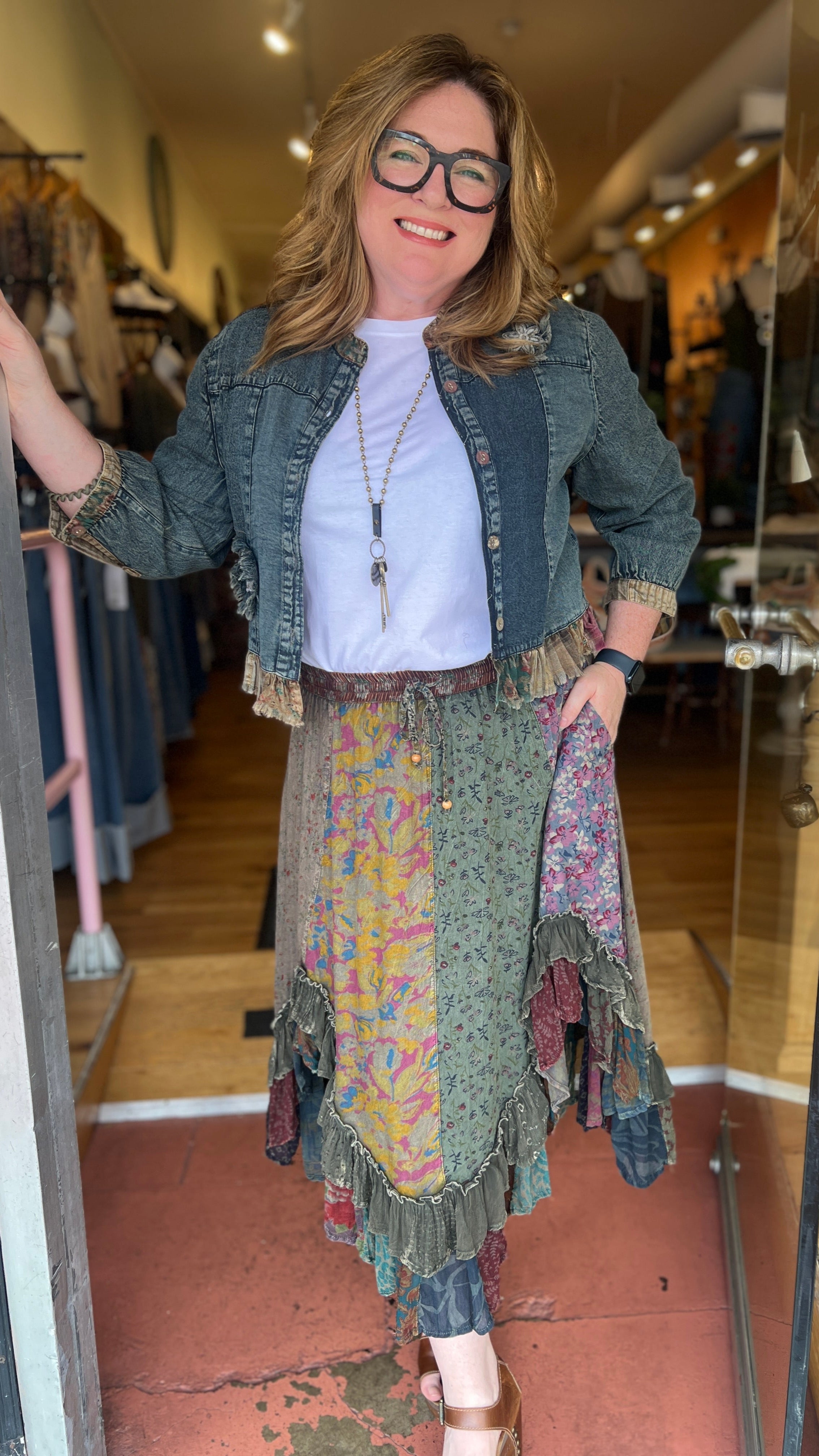 Woman standing in a store doorway wearing a patterned dress and denim jacket.
