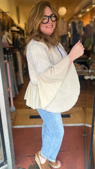 Woman standing in a store doorway wearing a beige cardigan, white top, blue jeans, and sandals.