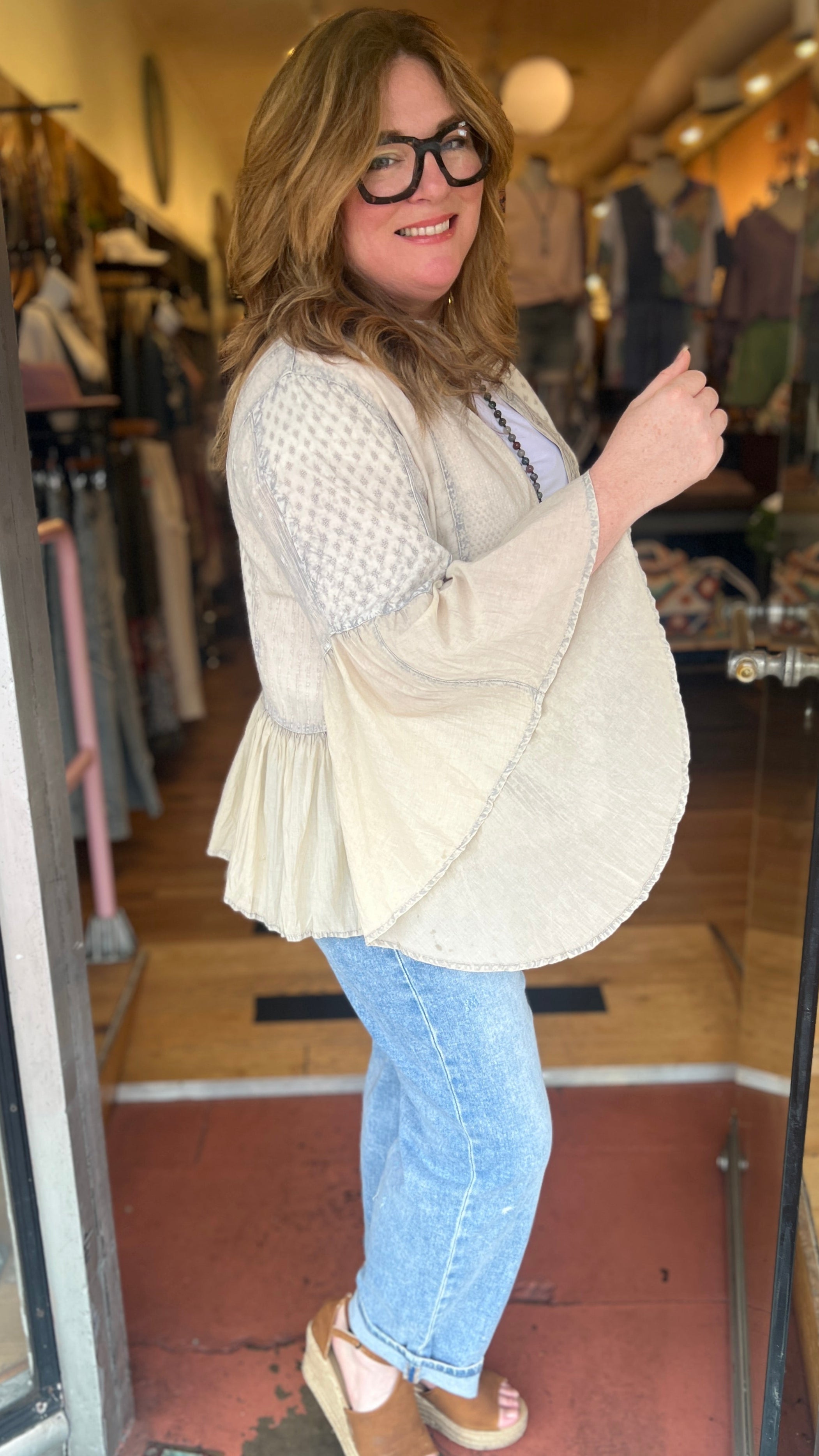 Woman standing in a store doorway wearing a beige cardigan, white top, blue jeans, and sandals.