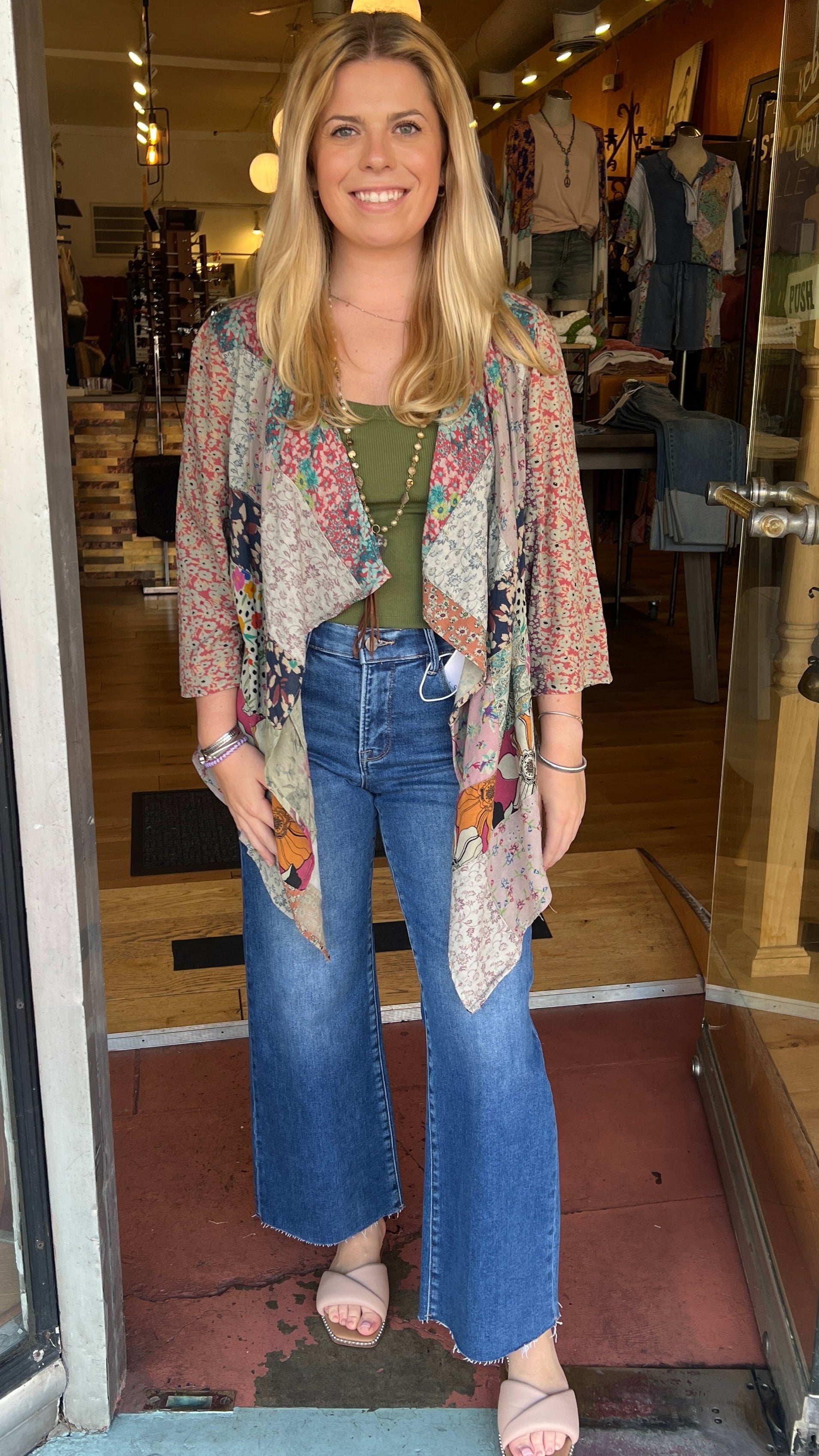 Woman standing in a store doorway wearing a floral kimono, green top, and blue jeans.