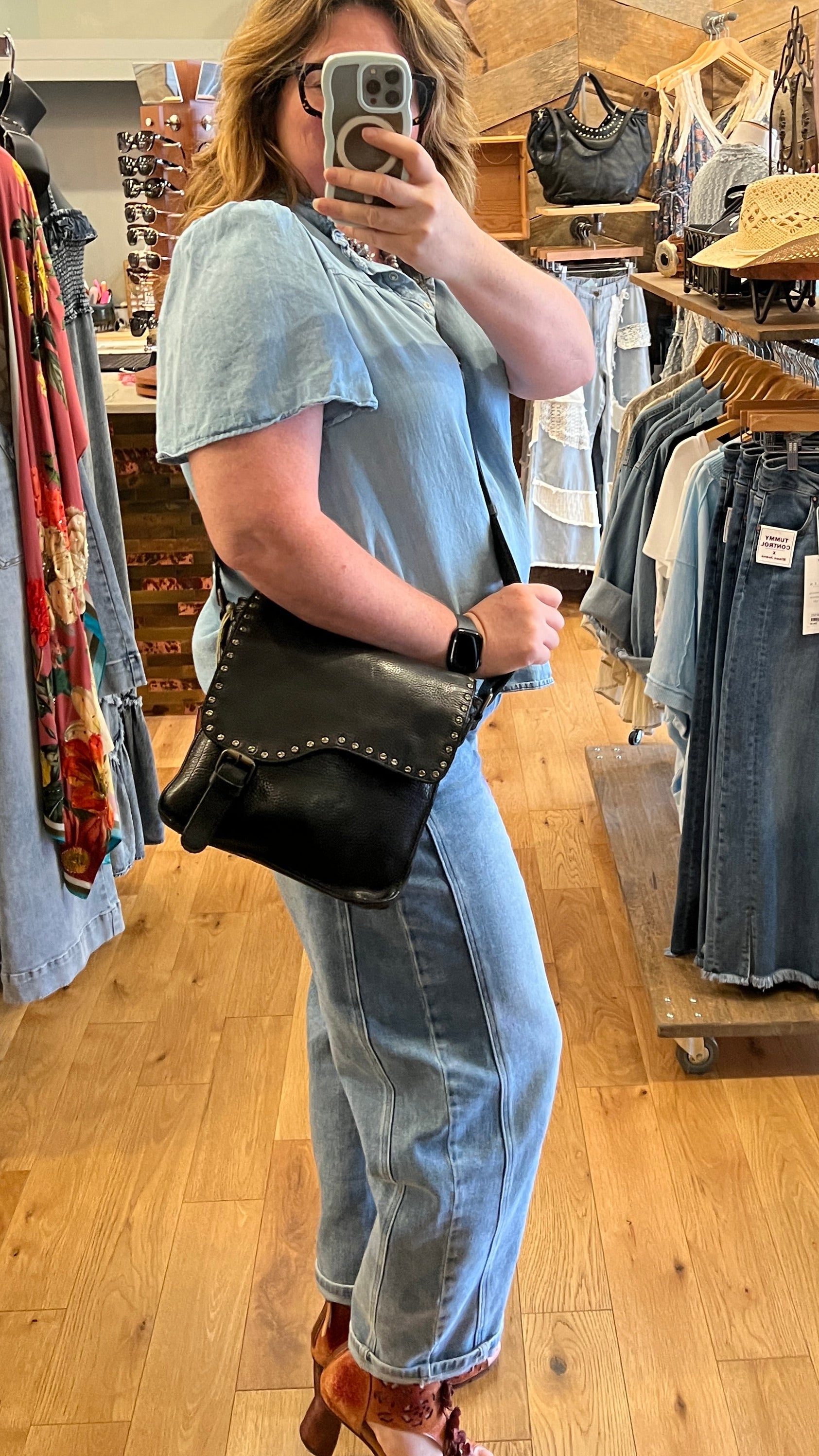 Person taking a mirror selfie in a clothing store with jeans displayed on racks.