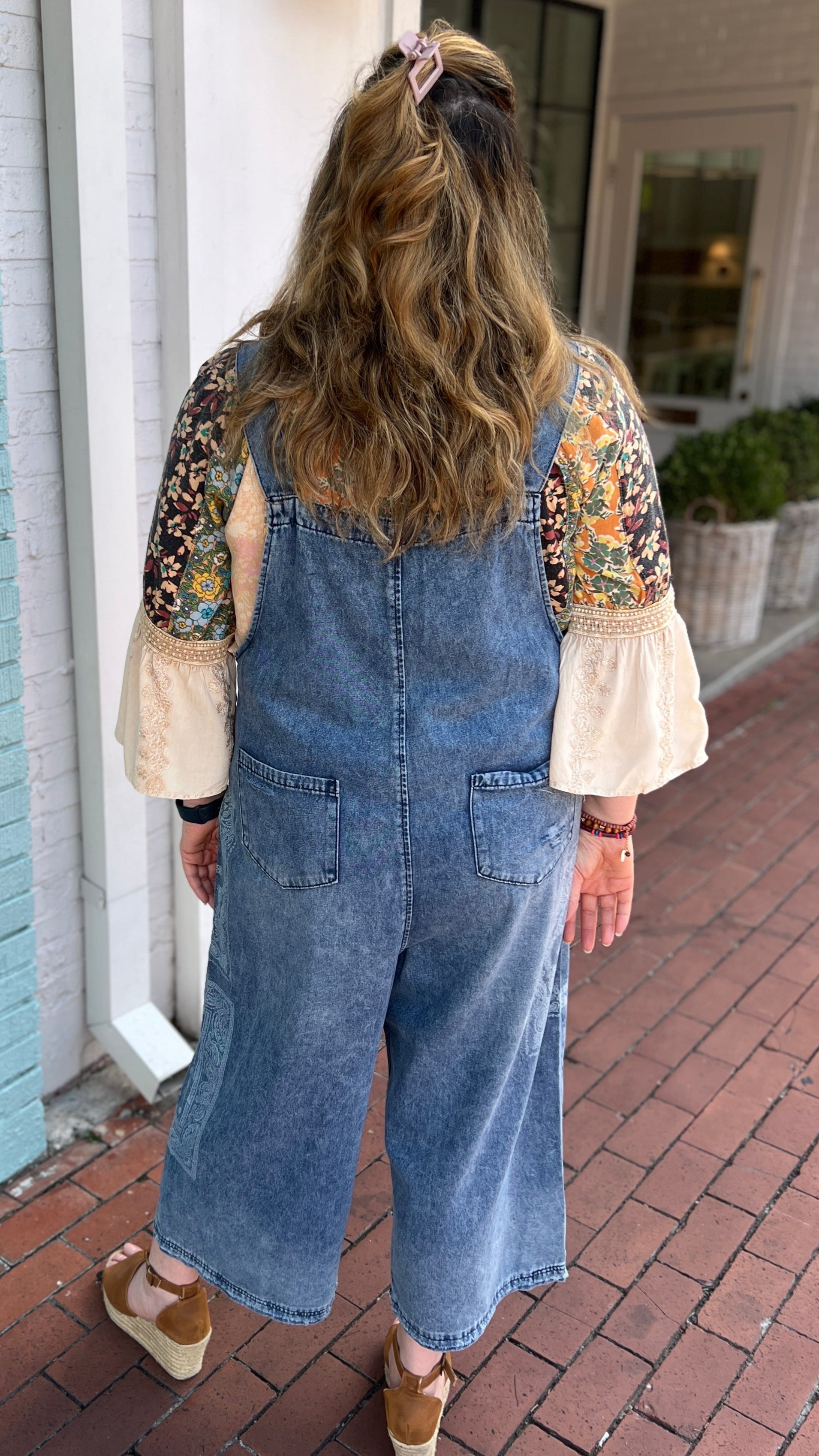 Person wearing denim overalls standing in front of a light blue brick wall.