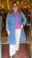 Woman wearing a long denim coat with floral embroidery, standing in front of a store entrance.