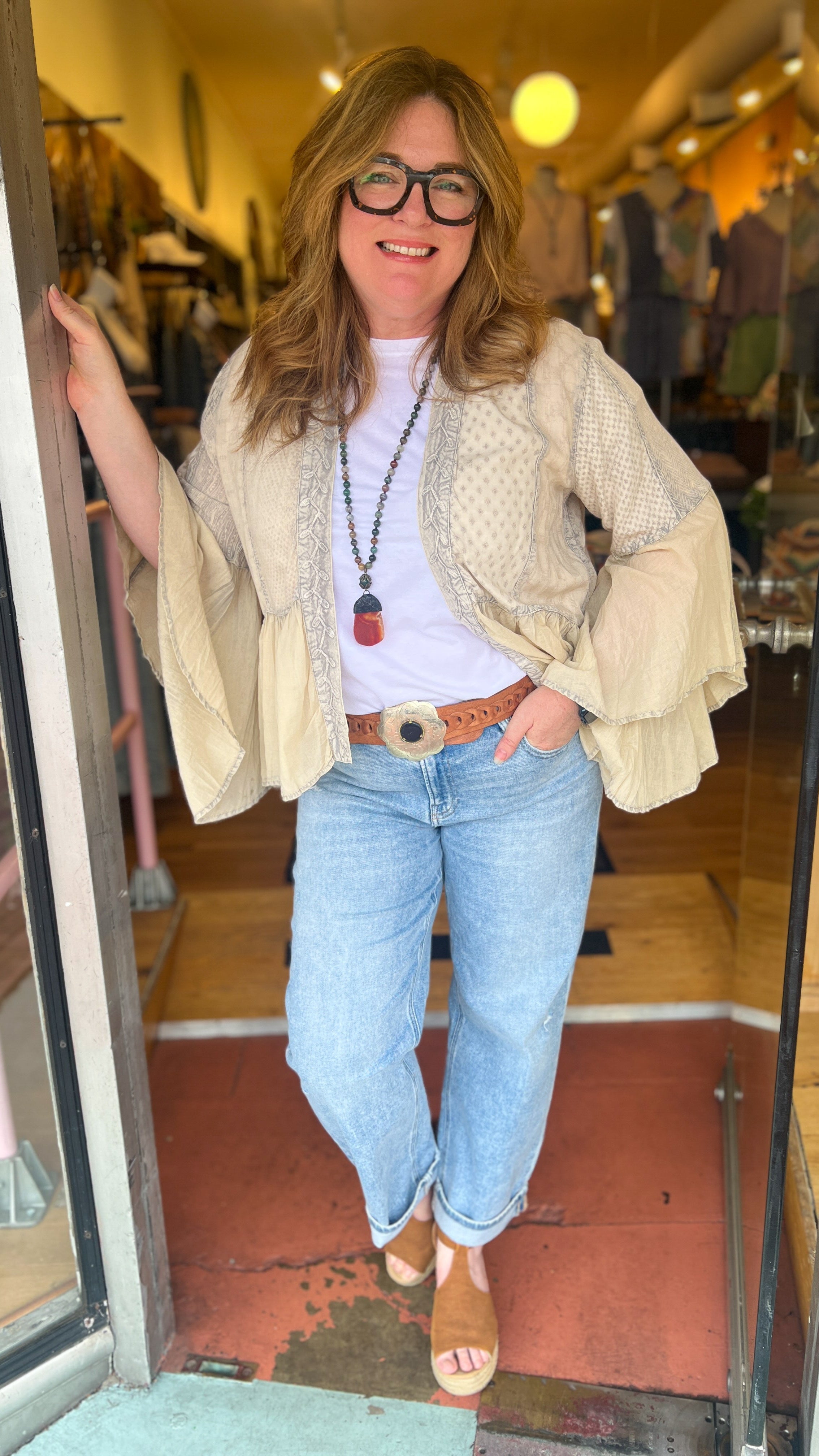 Woman standing in a store doorway wearing a beige jacket, white shirt, blue jeans, and glasses.