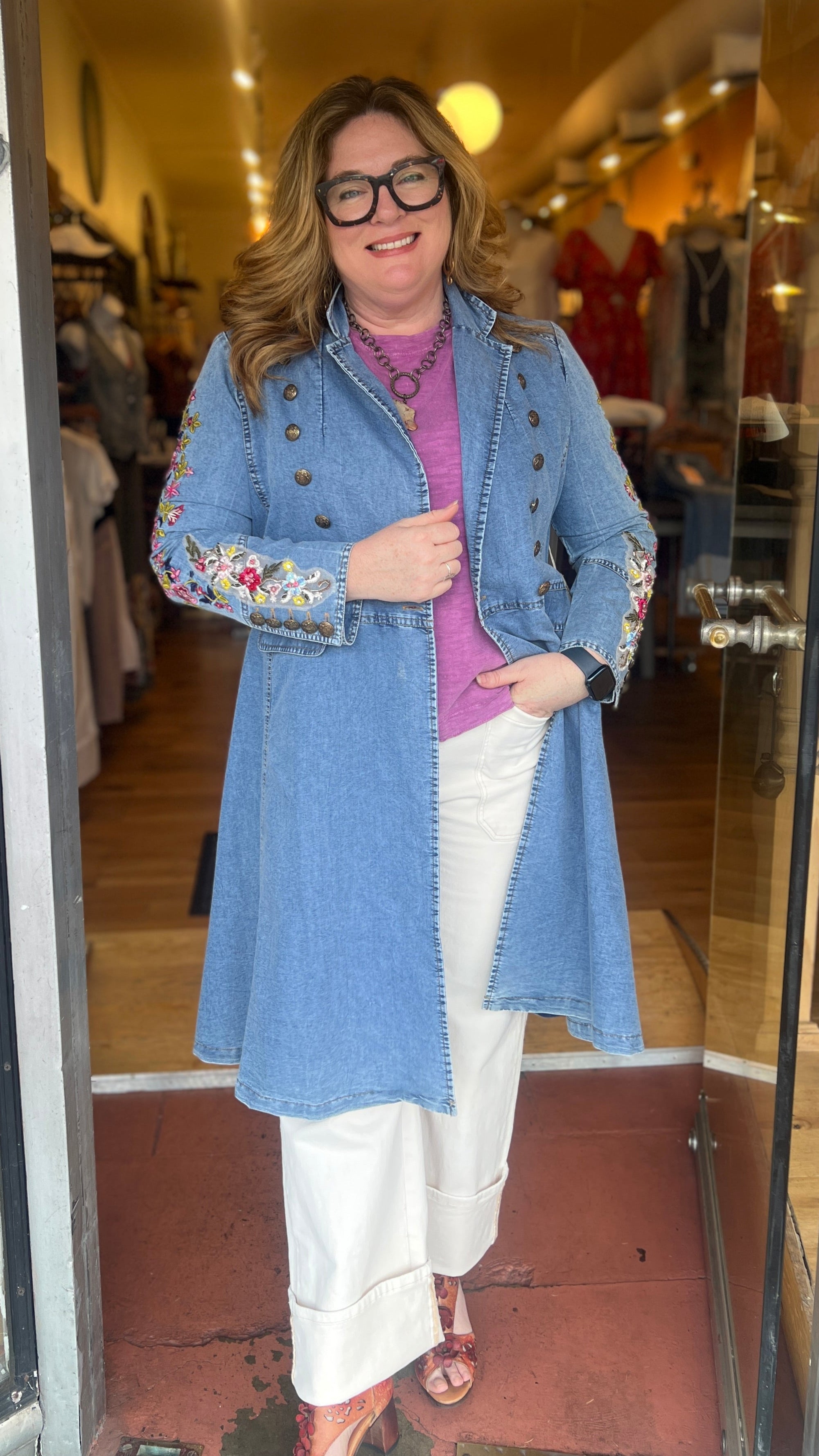 Woman wearing a blue coat with floral embroidery in front of a store entrance.