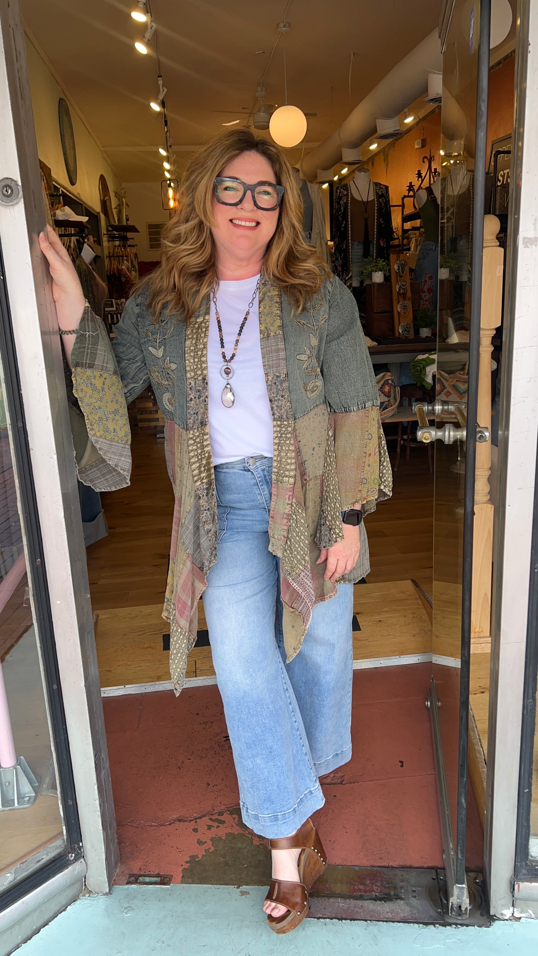 Woman standing in a store doorway wearing a green cardigan, white shirt, and blue jeans.