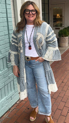 Person wearing a patterned cardigan and jeans standing on a sidewalk next to a brick wall.