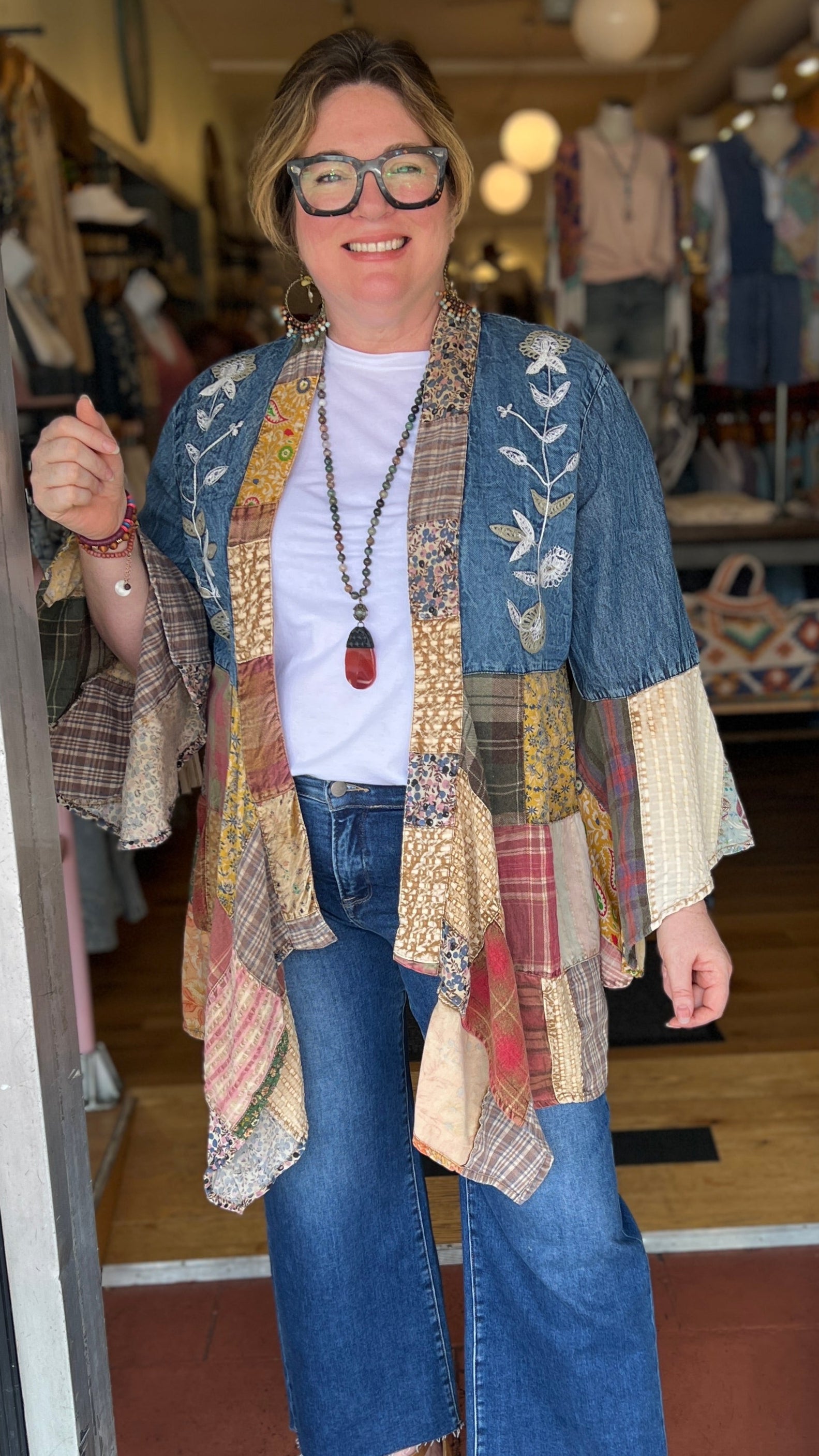 Woman standing in a store doorway wearing a patchwork cardigan and blue jeans.