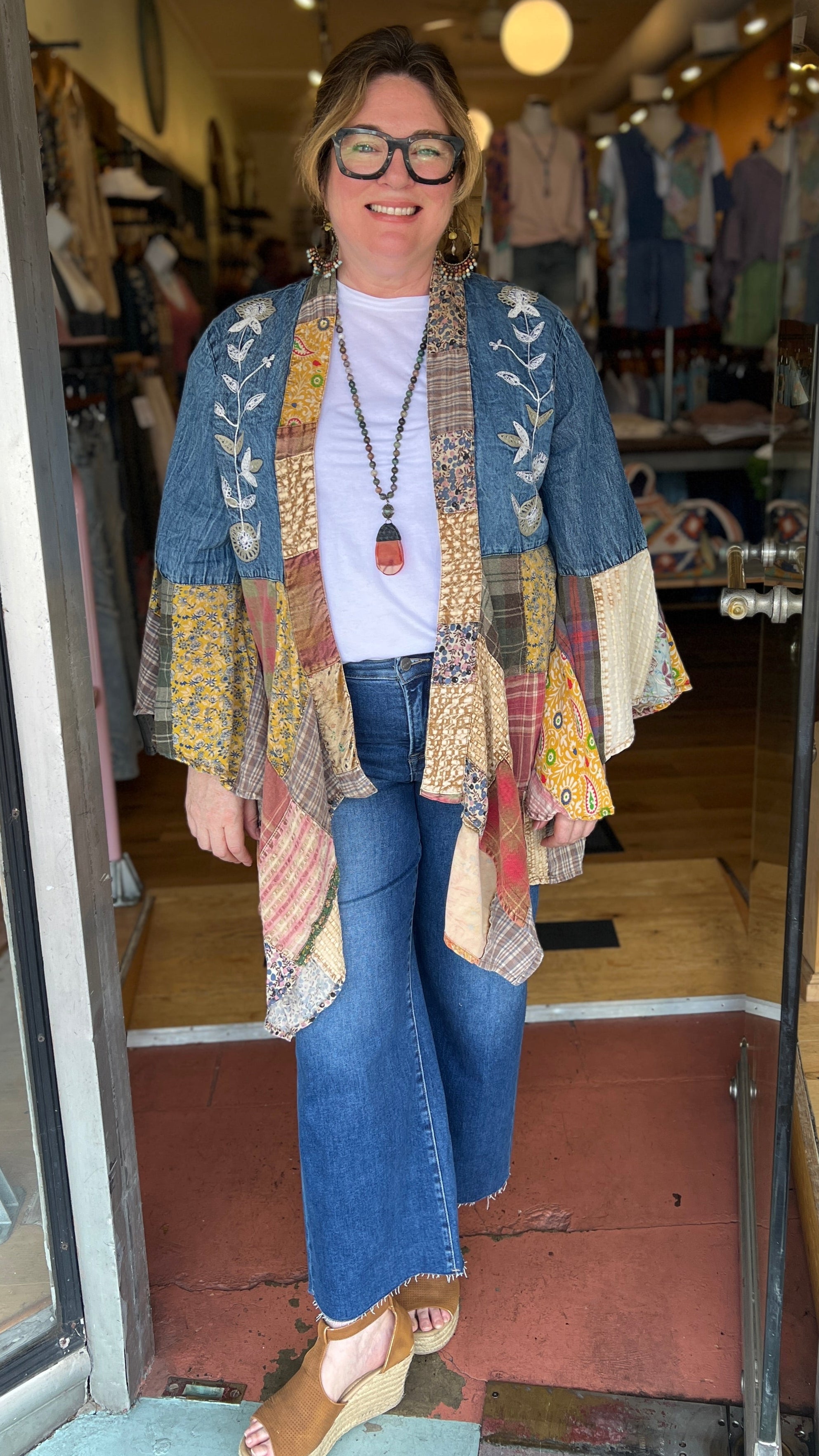 Woman standing in a store doorway wearing a colorful cardigan and blue jeans.