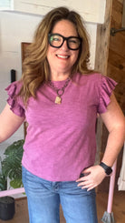 Person wearing a purple shirt and blue jeans standing in a store with shelves and products in the background.
