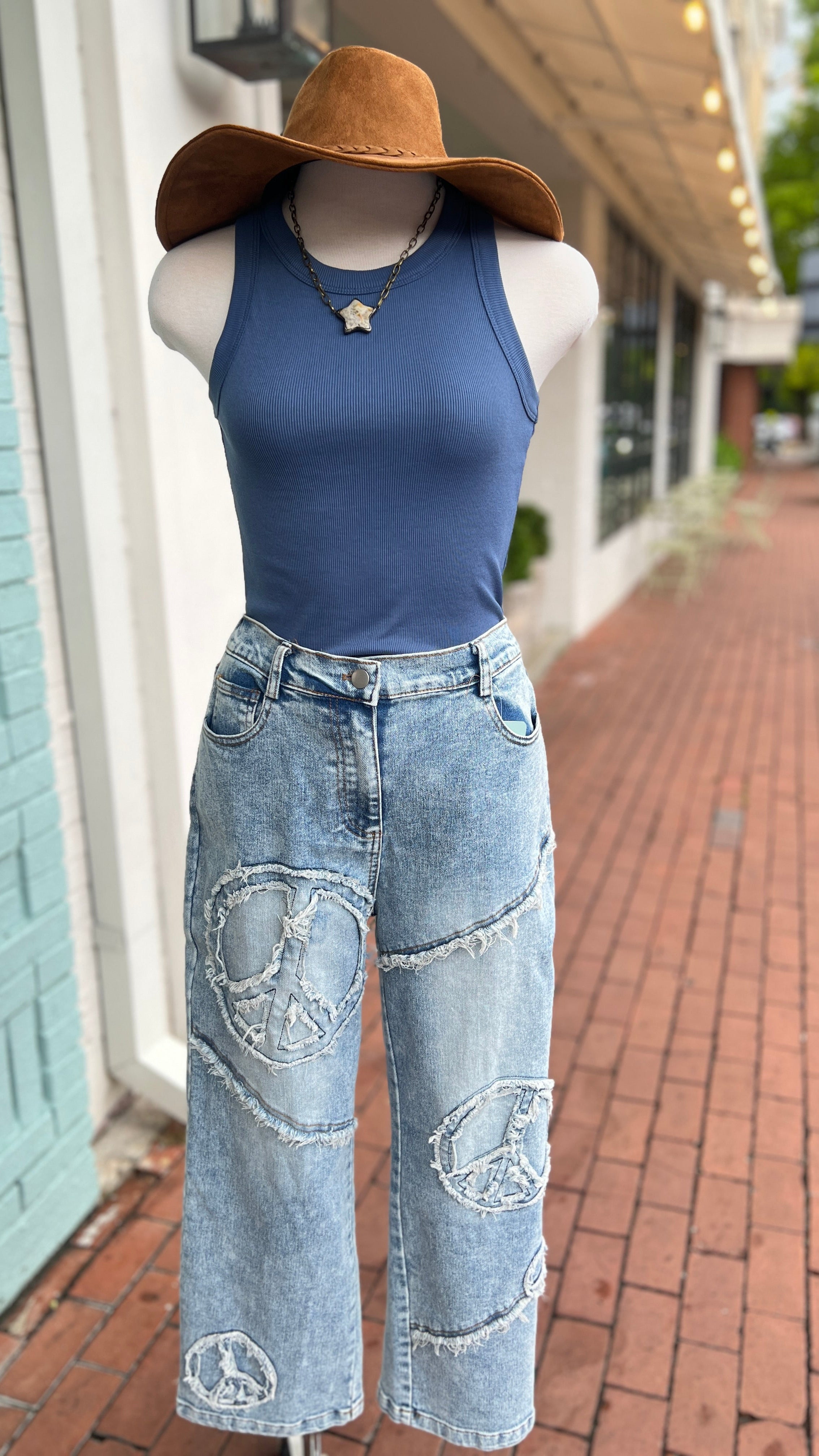 A mannequin wearing light wash denim jeans with a peace sign applique and a blue ribbed tank top, accessorized with a brown hat and necklace.