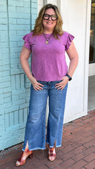 Woman in a purple shirt and blue jeans standing on a sidewalk in front of a building.
